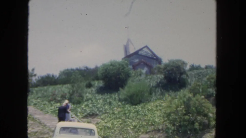 1962 San Diego 8mm frame showing visitors near a white car below a lush hillside and a glass-and-wood structure.