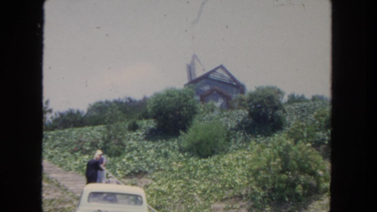 1962 San Diego home-movie view of two visitors by a white car below a modern hillside building and dense greenery.