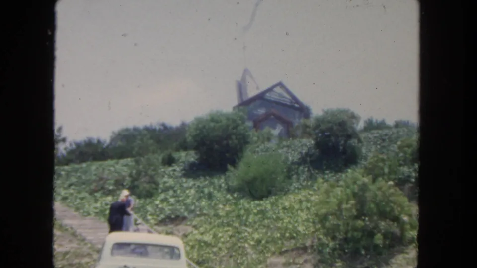 1962 San Diego home-movie view of two visitors by a white car below a modern hillside building and dense greenery.
