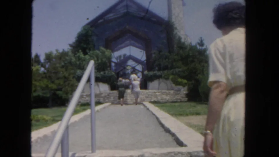 1962 San Diego home-movie frame showing a visitor on a paved path with railings leading to a hillside glass-and-wood building.