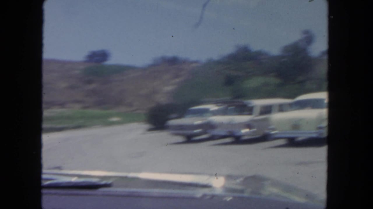 Classic cars parked along a curved hillside road seen from inside a vehicle.