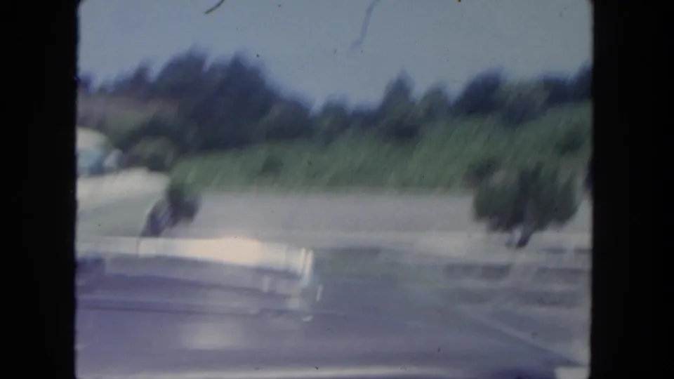 Motion-blurred 1962 San Diego 8mm driving view with roadside shrubs, green hillside, pavement, and the filming car hood visible.