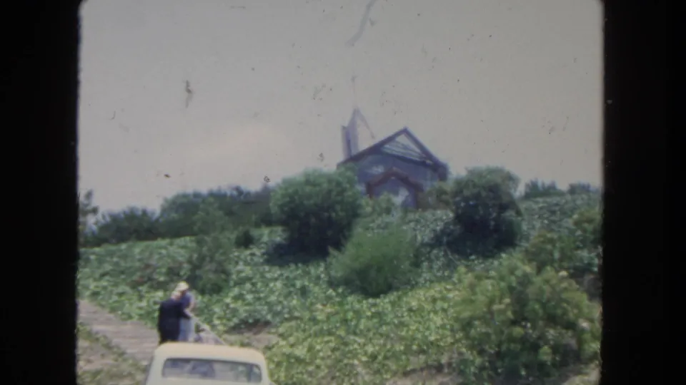1962 San Diego home-movie frame with a person by a light-colored car, dense hillside planting, shrubs, and a peaked-roof structure above.