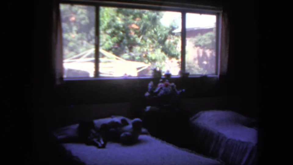 1962 San Diego home-movie frame of a dim bedroom with twin beds and a bright garden or tree view through the window.