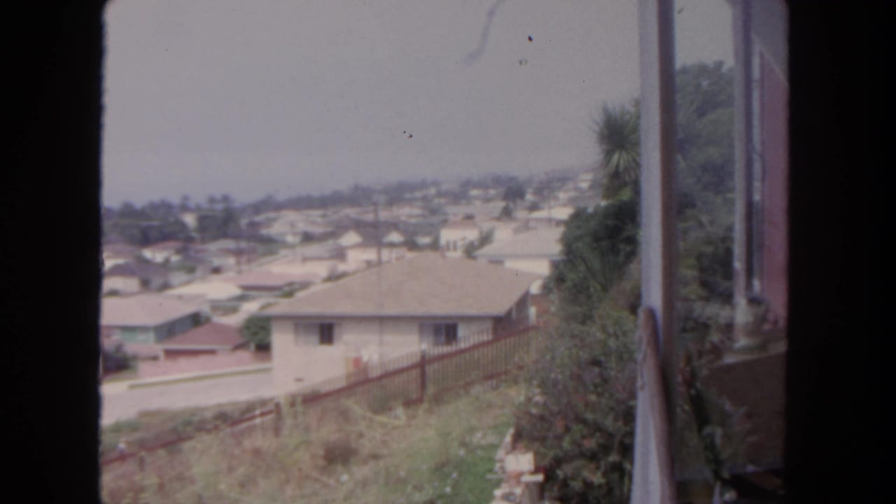 Hillside suburban houses seen through a window with a street and fence below.