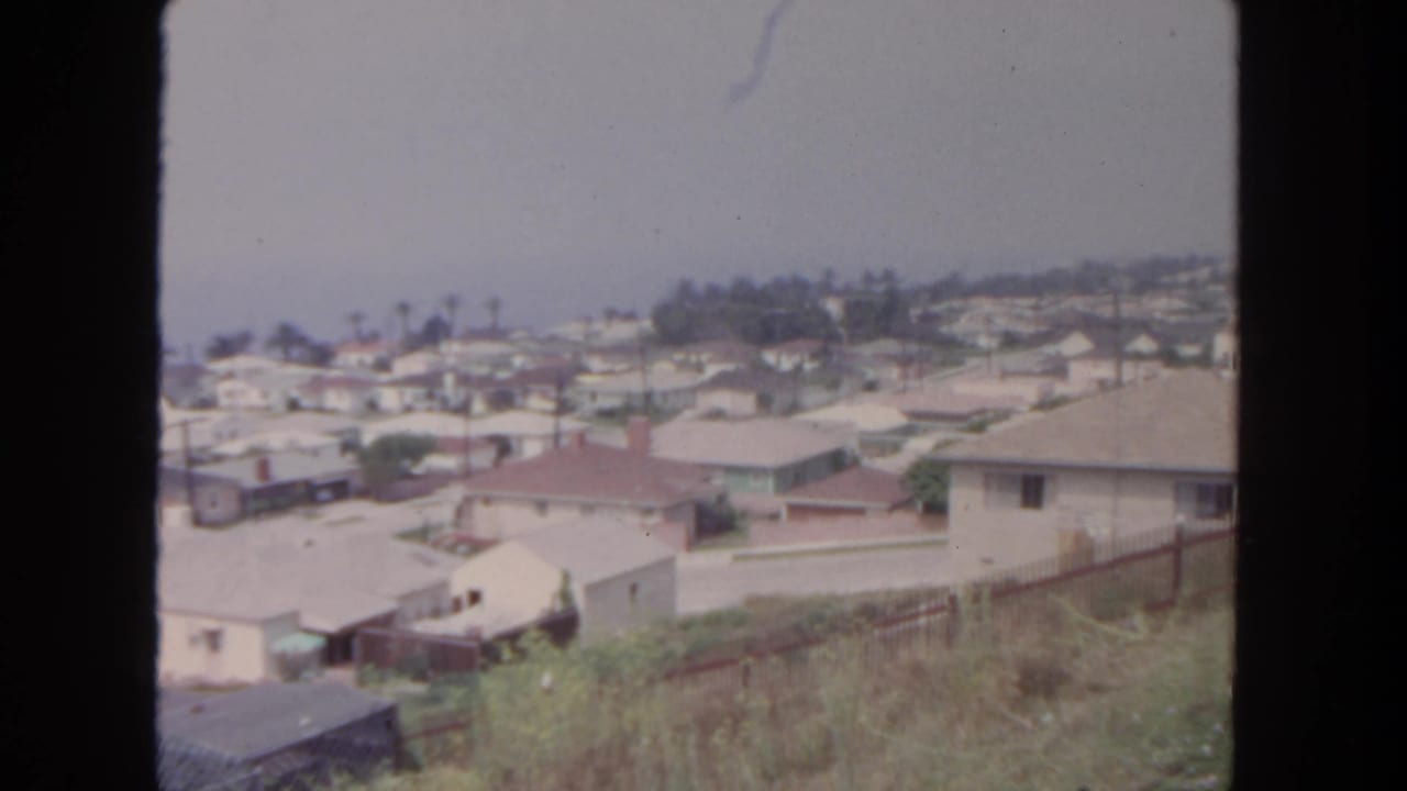 Elevated view across a residential neighborhood with rooftops, fences, brush, and hazy sky.