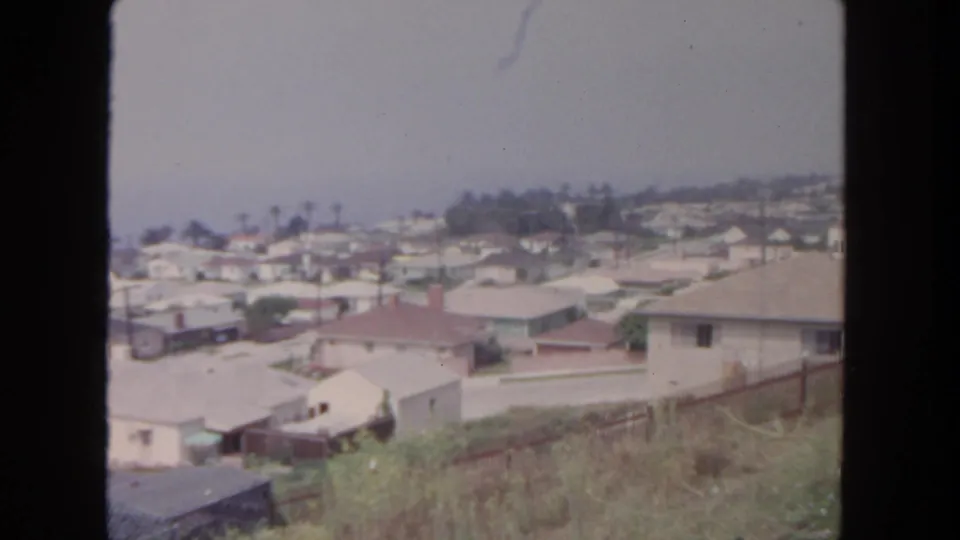 1962 San Diego home-movie frame looking over low-rise homes, fenced yards, hillside brush, and a hazy sky.