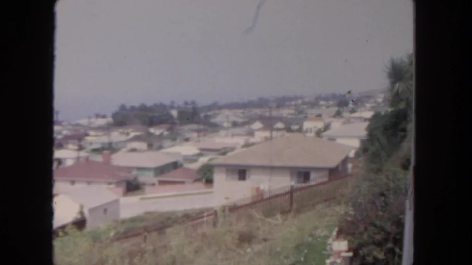 1962 San Diego 8mm frame showing a hillside vantage over homes, fence lines, vegetation, and a hazy residential background.