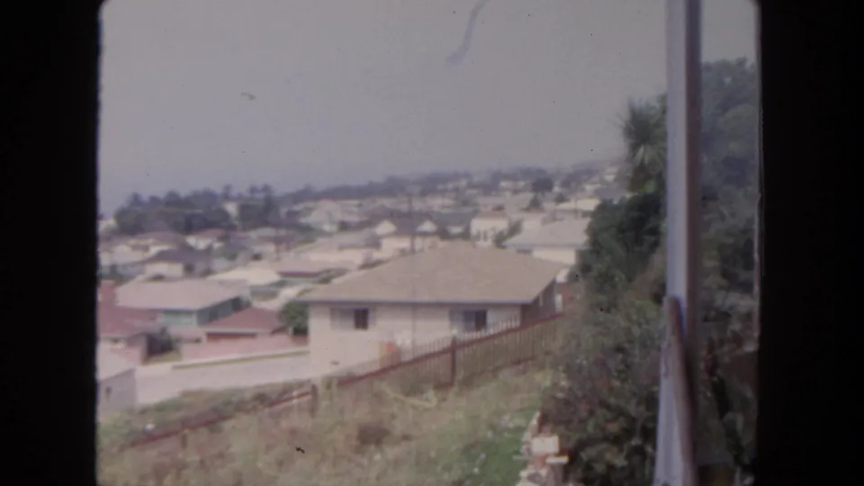 1962 San Diego home-movie frame at the end of the pan, showing rooftops, hillside brush, trees, and a vertical structure at right.