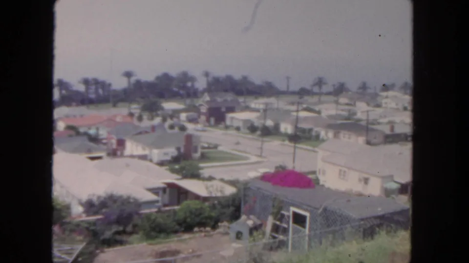 1962 San Diego home-movie frame showing an elevated suburban view with tract houses, pale rooftops, palms, streets, lawns, and a vivid pink shrub.