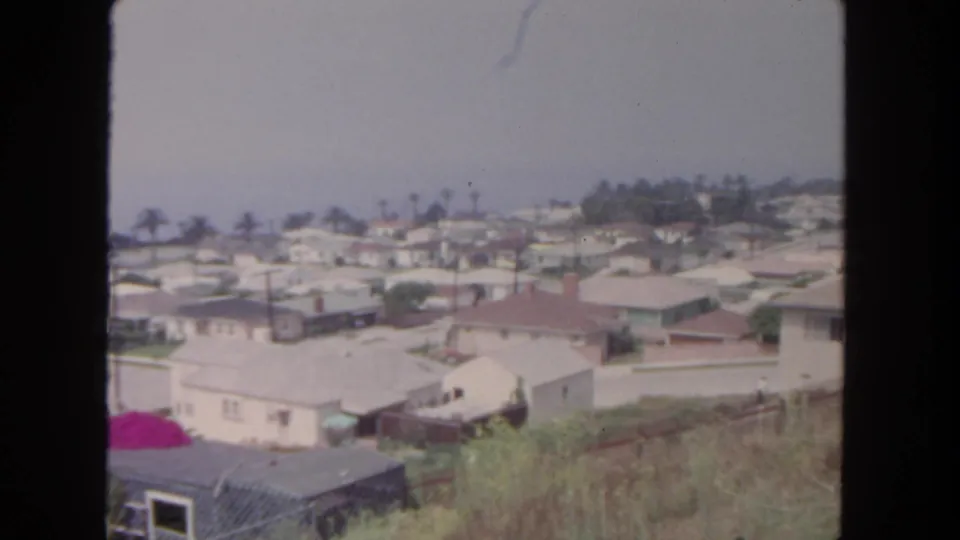 1962 San Diego home-movie view over dry hillside grass toward rows of single-story houses, pale roofs, streets, palms, and utility poles.
