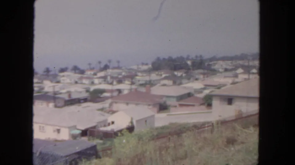 1962 San Diego frame showing a wide elevated residential panorama with single-story houses, pale rooftops, palms, utility poles, and dry foreground grass.