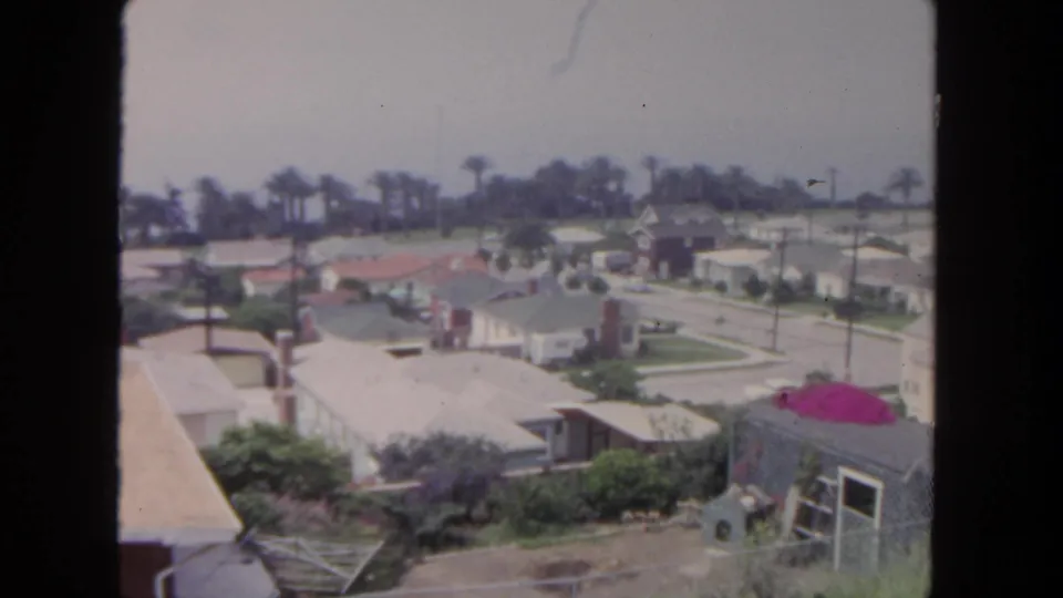1962 San Diego home-movie view across suburban rooftops, palms, streets, and a small outbuilding with a bright magenta roof area.