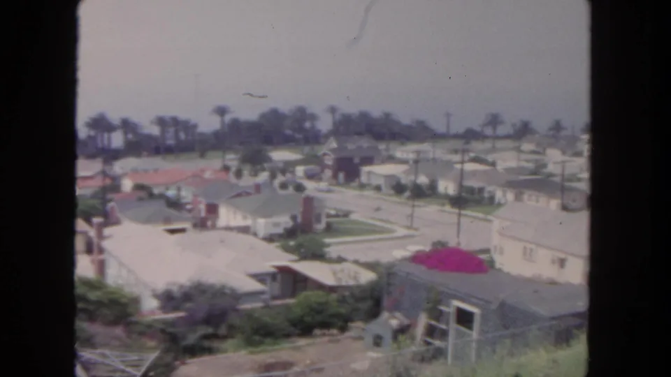 1962 San Diego frame showing low suburban houses, lawns, palm trees, utility poles, and a small outbuilding from above.