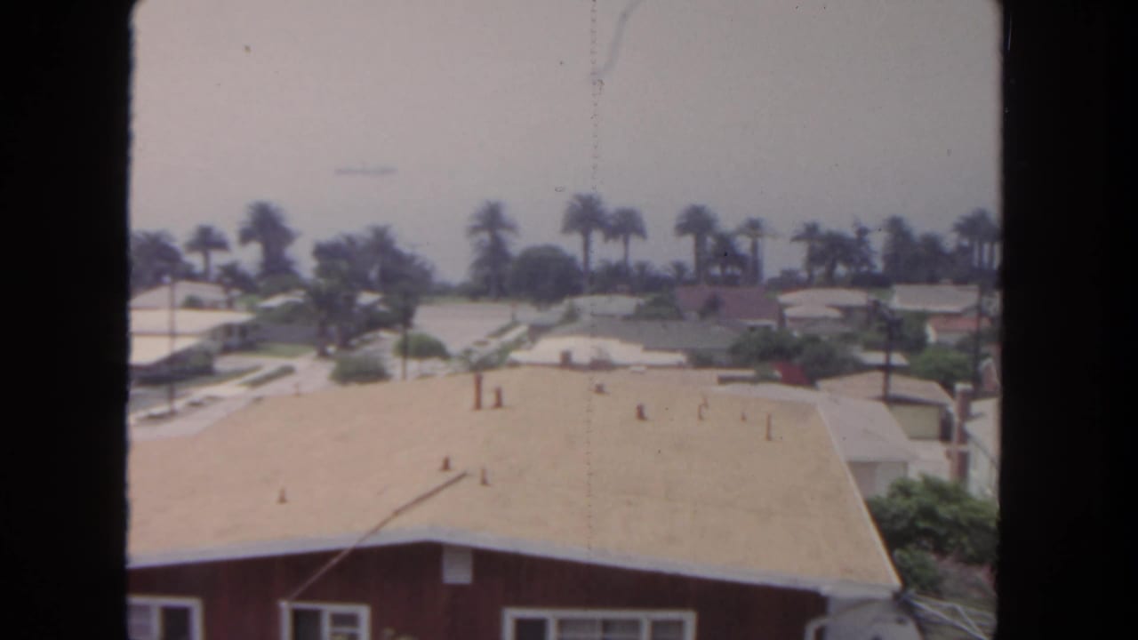 1962 San Diego home-movie frame looking over a pale roof toward palm trees, lawns, and low suburban homes.
