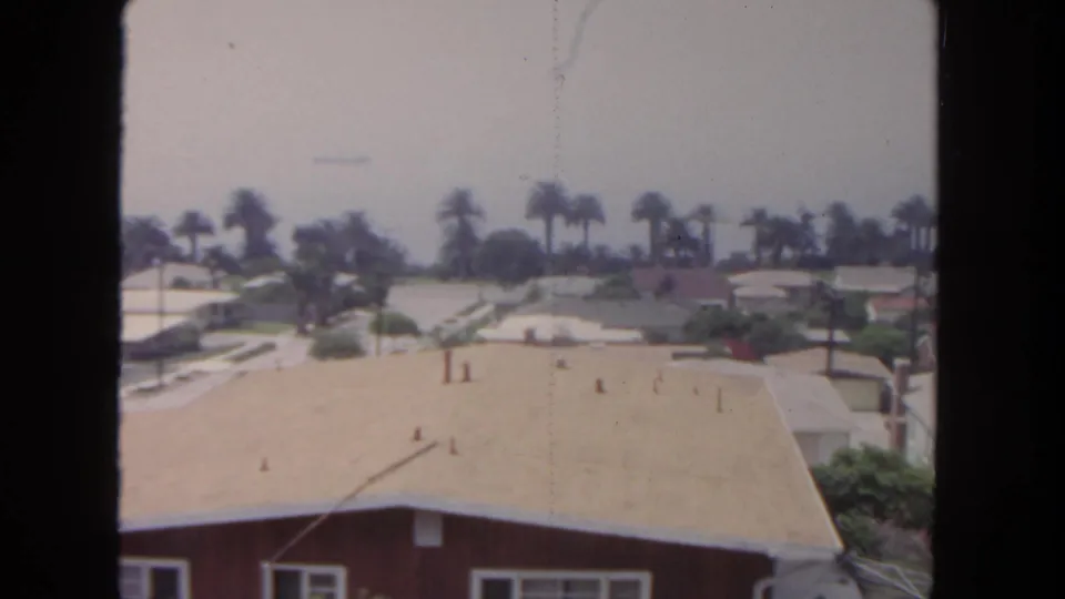 1962 San Diego home-movie frame looking over a pale roof toward palm trees, lawns, and low suburban homes.