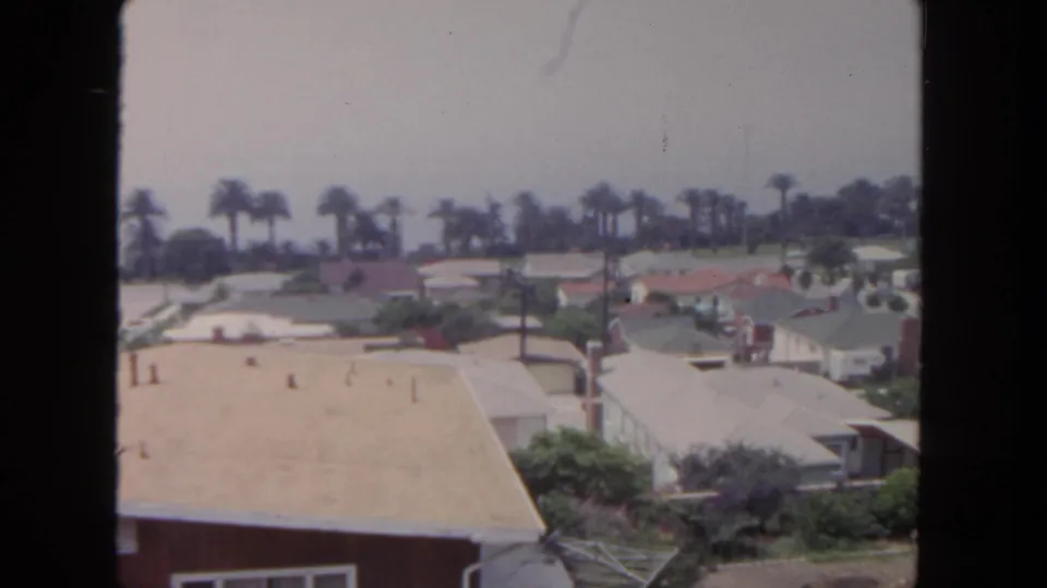 1962 San Diego frame of a quiet suburban block with pale rooftops, palms, driveways, shrubs, and utility lines.