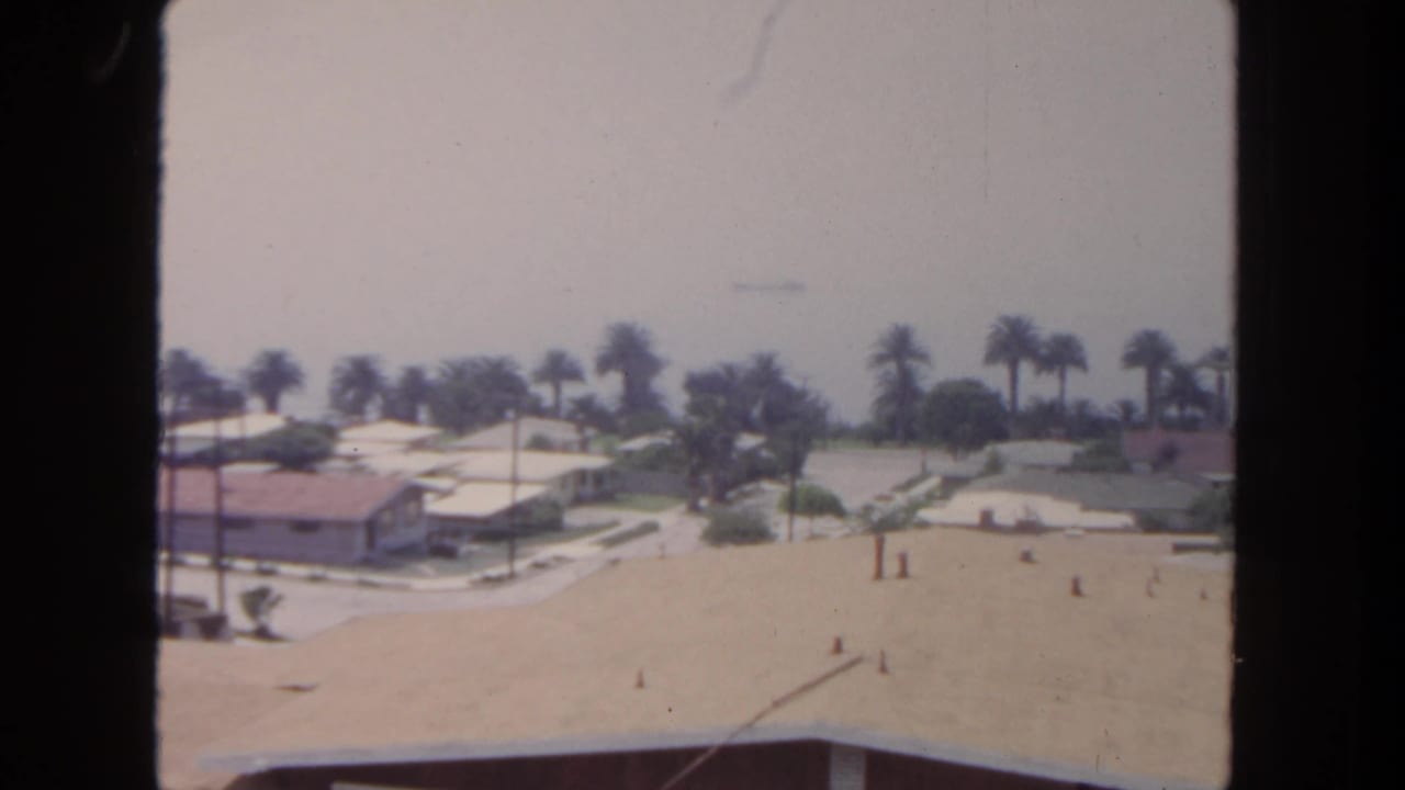 1962 San Diego suburban view with palm trees, low residential buildings, pale roofs, and a soft hazy horizon in 8mm color.