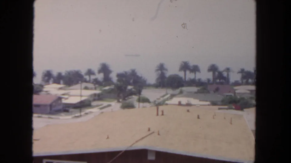 Elevated 1962 San Diego home-movie frame showing rooftops, palms, a quiet street grid, and a hazy coastal-looking background.