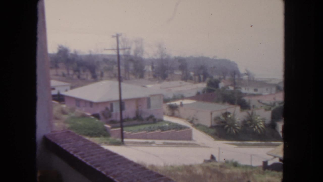 Suburban street seen from above with single-story homes, garden walls, utility poles, and distant haze.