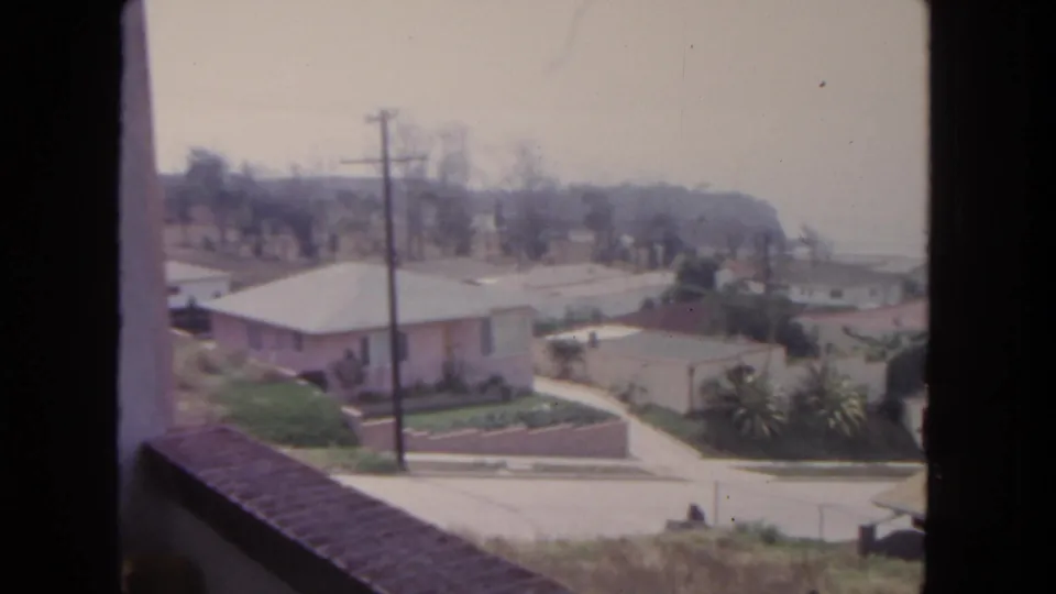 1962 San Diego suburban neighborhood viewed from an elevated window, with houses, palms, utility poles, and a hazy hillside beyond.