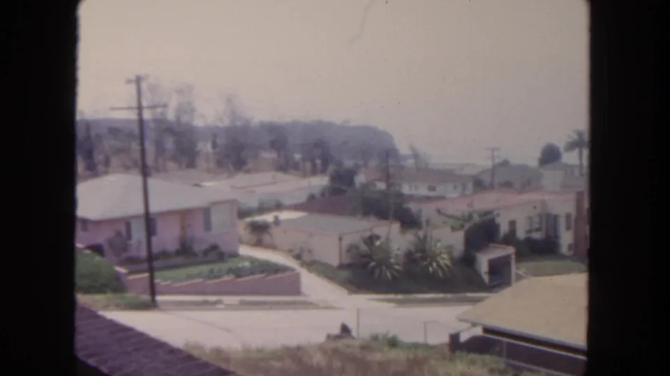 1962 San Diego street view showing low suburban homes, landscaped yards, utility poles, palms, and a soft hazy sky.