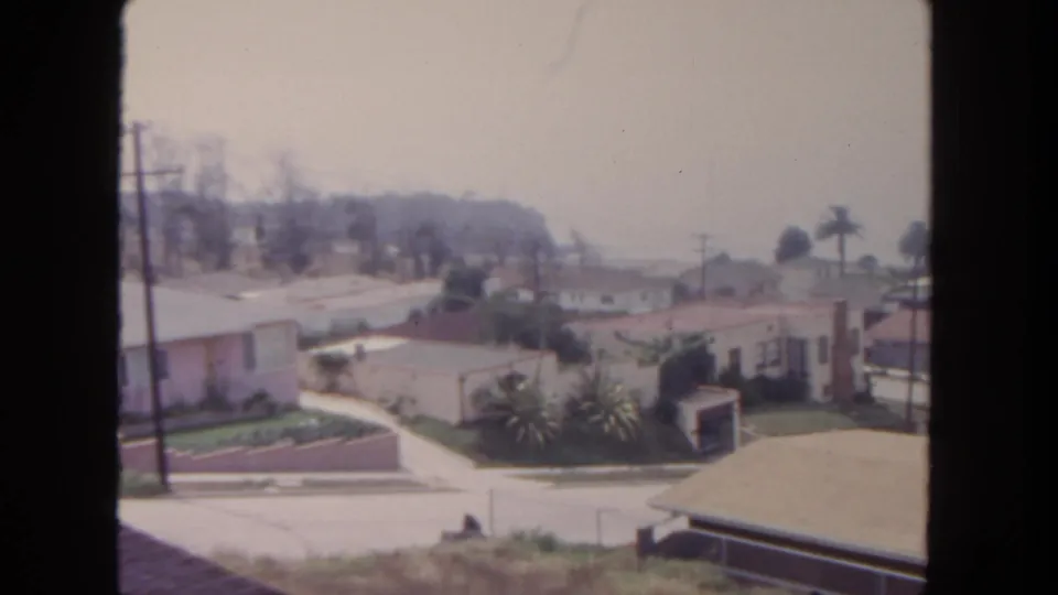 1962 San Diego neighborhood view with tile-roof houses, palms, low garden walls, a curving street, and distant residential slopes.