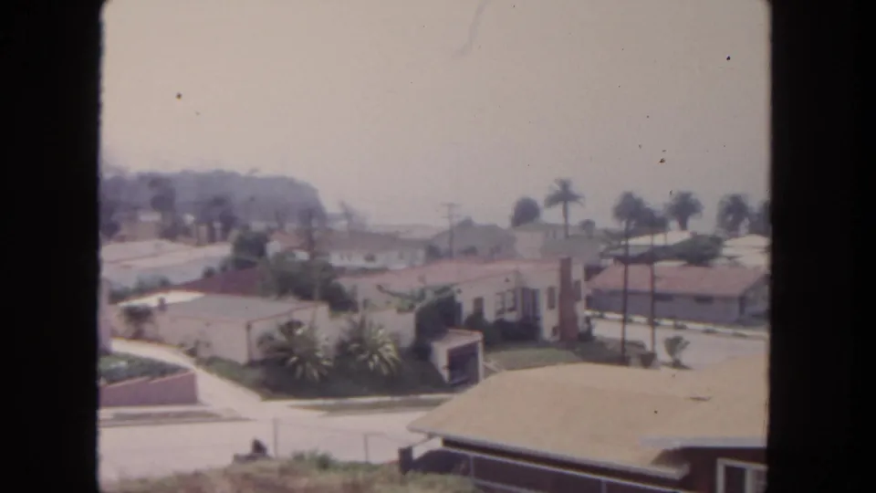 1962 San Diego suburban houses and palms seen across a quiet street, with utility lines, roof edges, and a hazy hillside behind.