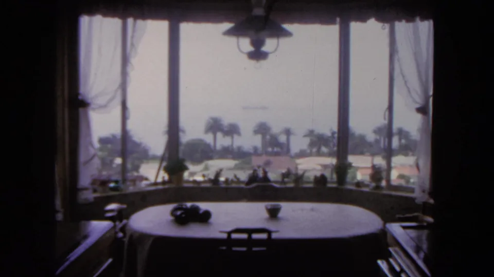 1962 San Diego home-movie view from a dining room table through a bay window toward palms, hazy water, and a distant ship.