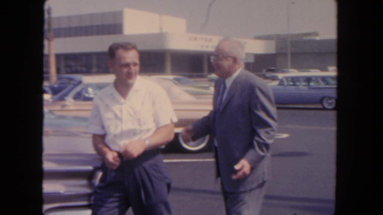 In 1962 San Diego footage, two adult men appear to finish a conversation beside parked early-1960s cars near a terminal-like building.