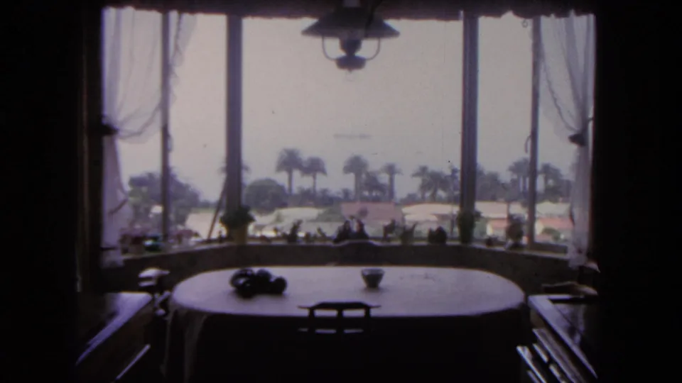 The home-movie view lingers on a windowed San Diego interior with a round table, dark furnishings, palm trees, and a bright exterior beyond.