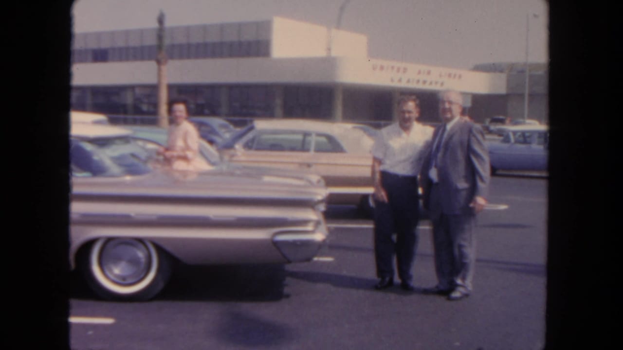 Two men stand near a silver sedan in an airport parking lot with a terminal behind them.