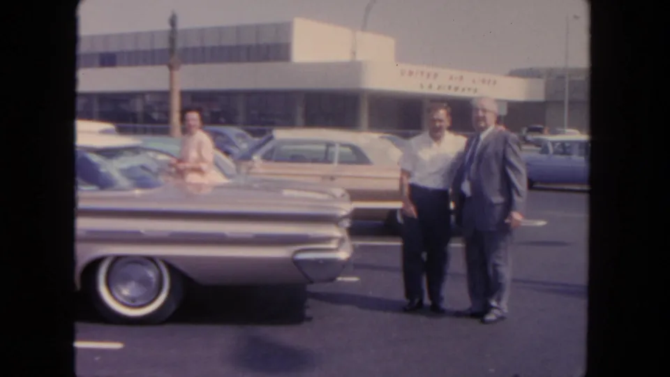 In San Diego in 1962, two adult men stand beside a silver sedan while a United Air Lines terminal and parked cars sit behind them.