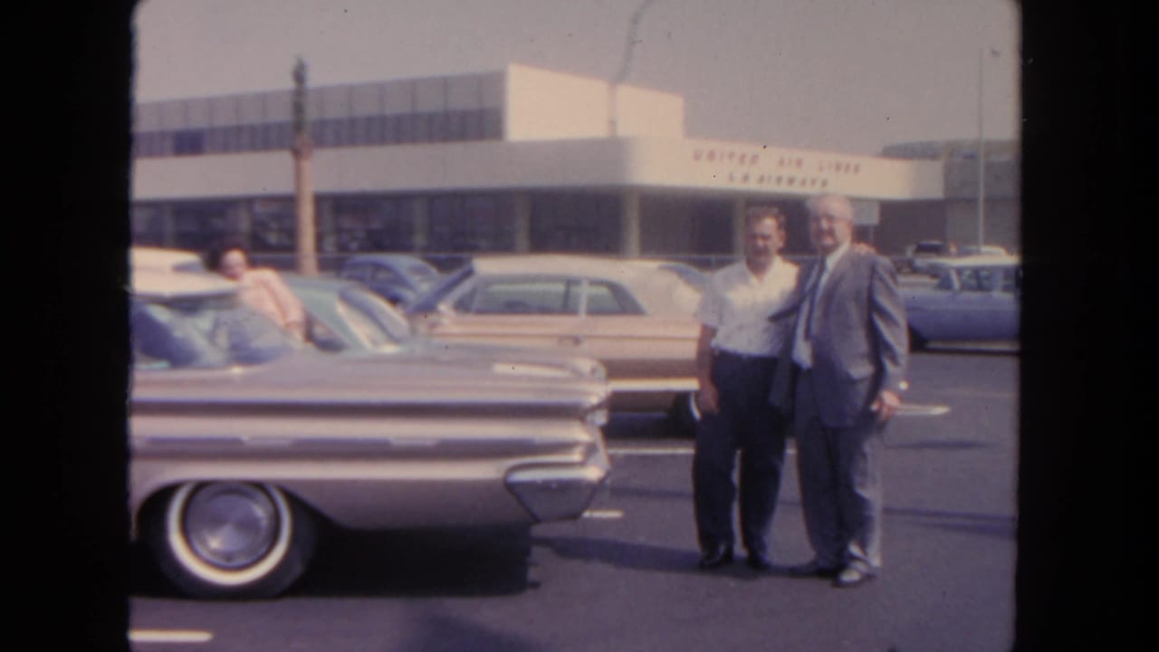Two men pose or talk in a 1962 San Diego airport parking lot, with a woman leaning into a car and airline signage in the background.