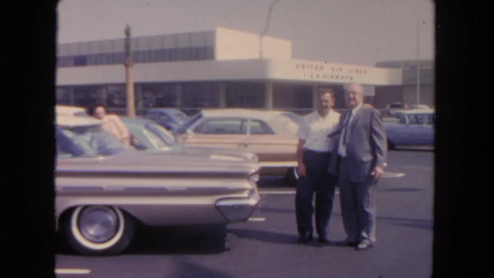 Two men pose or talk in a 1962 San Diego airport parking lot, with a woman leaning into a car and airline signage in the background.