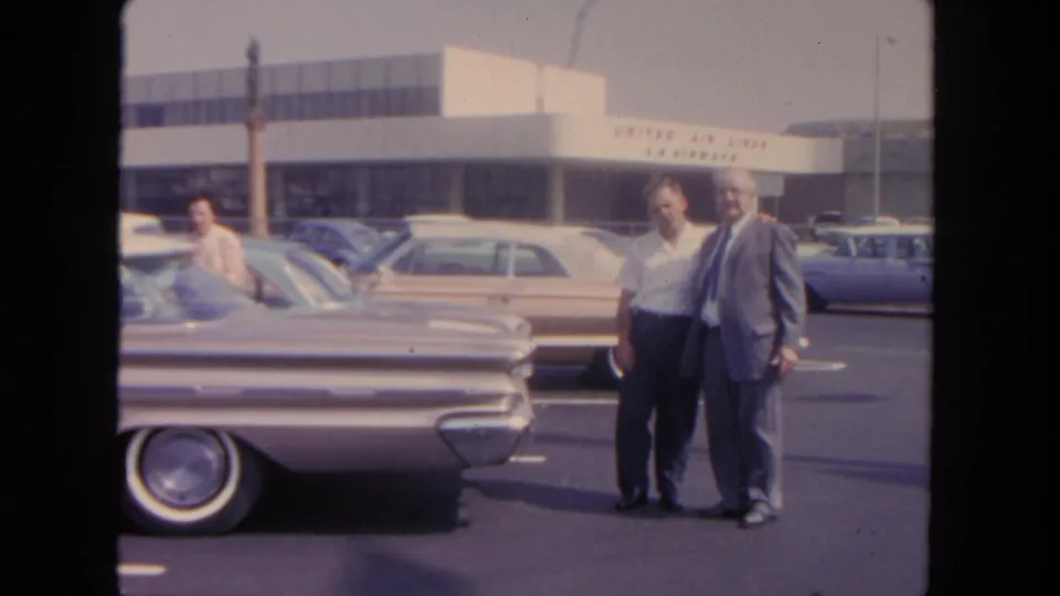 A 1962 San Diego airport parking lot view shows two men beside a silver sedan, with mid-century cars and a terminal facade behind them.