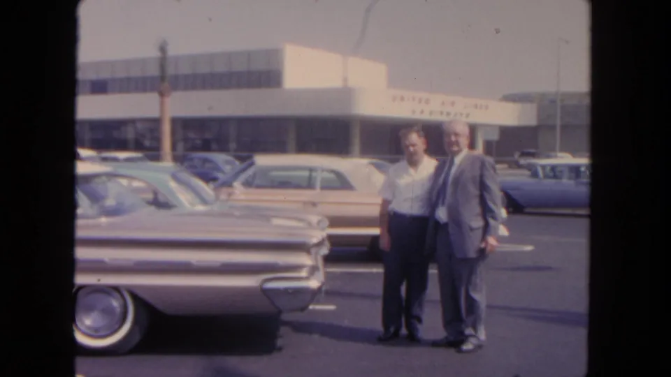 At the San Diego airport in 1962, two men stand near a classic sedan while parked cars and United Air Lines signage remain visible.