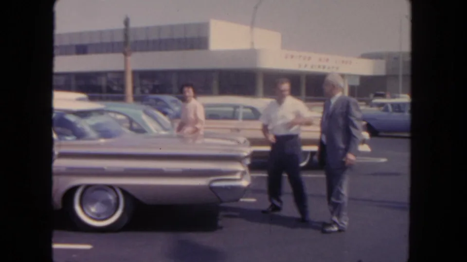 Ground-level 1962 San Diego airport scene with two men beside classic cars and visible United Air Lines terminal signage.