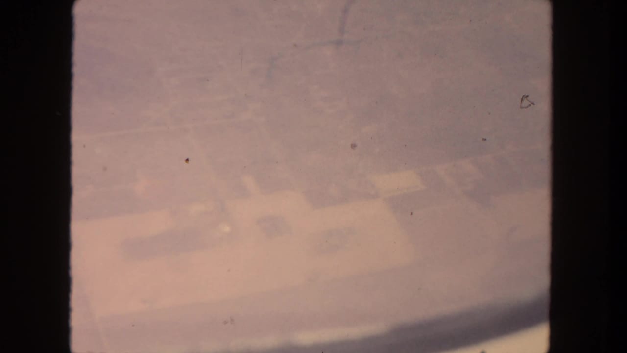 Aged 8mm aerial view over the San Diego area, with pale fields and faint settlement patterns under a pink color cast.
