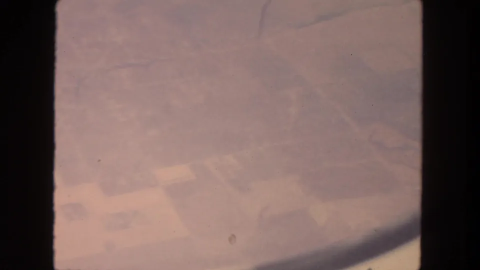 1962 aerial home-movie frame over farmland near San Diego, with blocky fields, small structures, and visible 8mm wear.