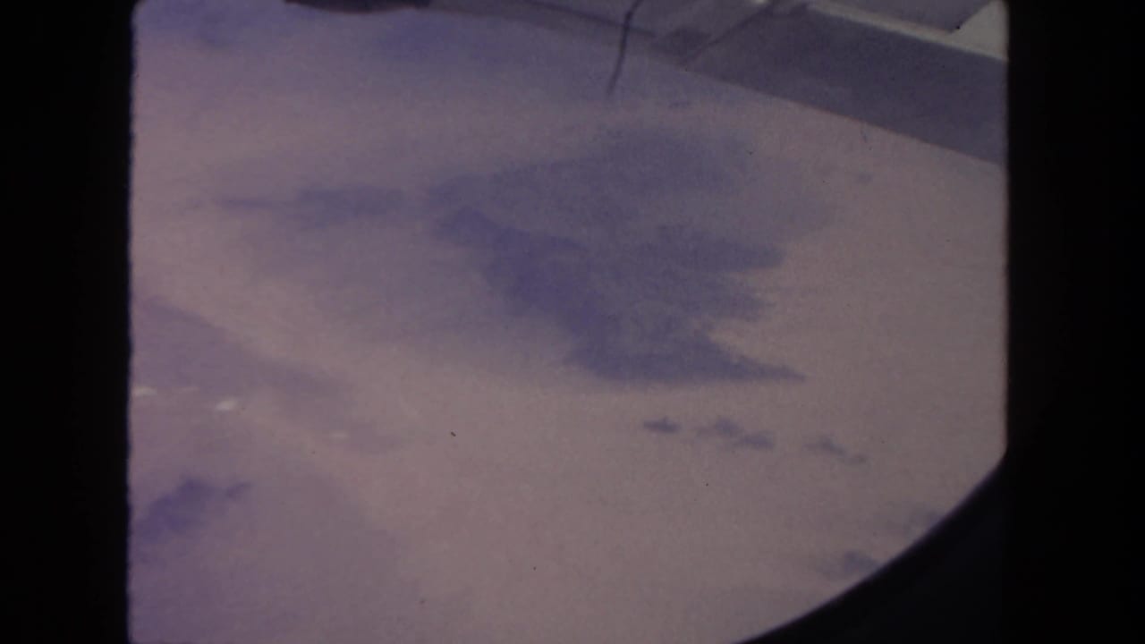 1962 8mm view from an airplane window, with indistinct terrain below and a dark rounded window edge.