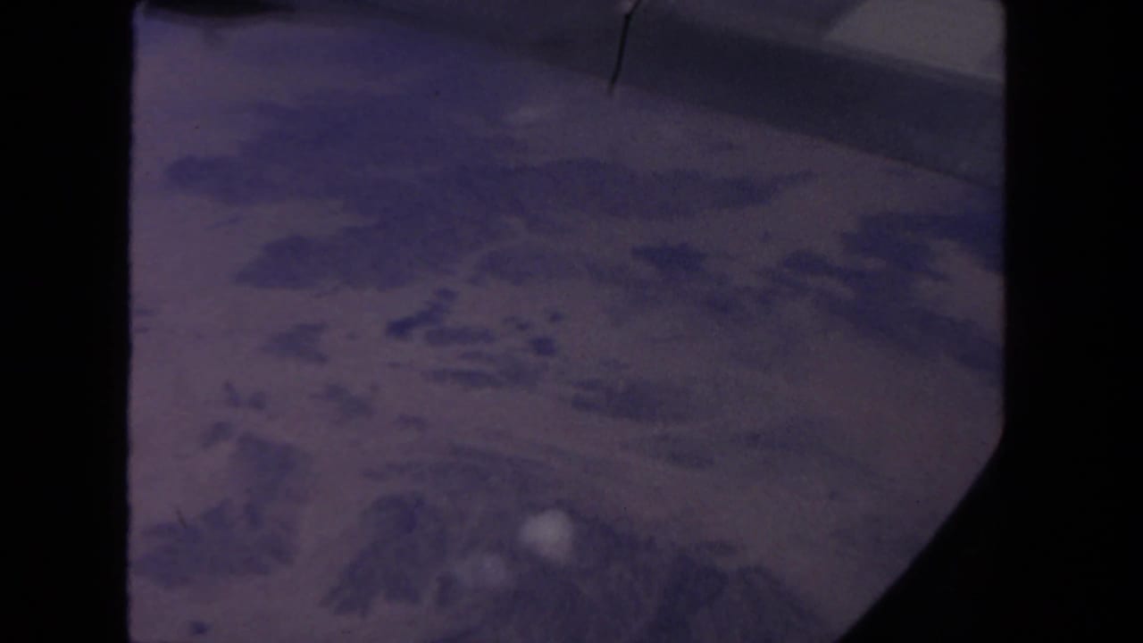 Hazy 1962 passenger-window aerial view with textured landforms and pale cloud patches below.
