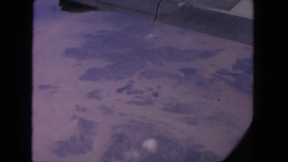 1962 8mm airplane-window view showing hazy land below and a small portion of aircraft structure above.