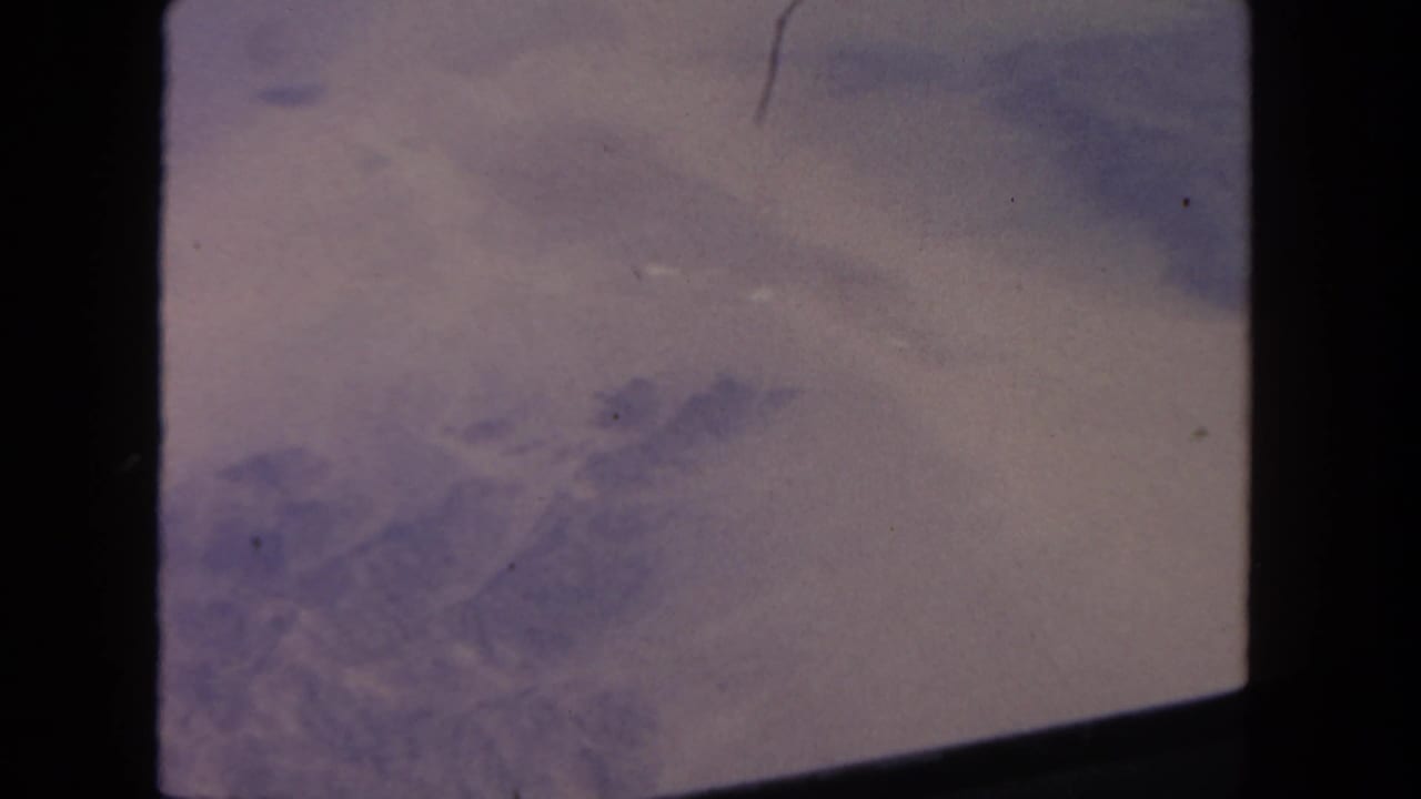 1962 8mm home-movie frame showing pale desert terrain and haze through an airplane window, with aged purple-beige color.