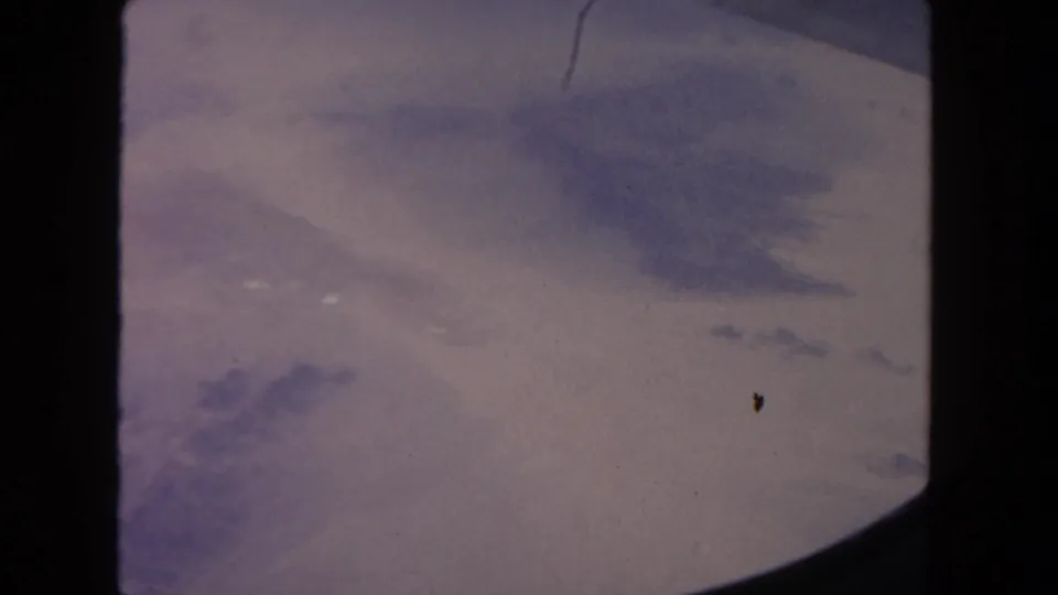 1962 8mm travel footage frame with soft arid landscape, cloud shadow, and dark airplane-window edges crossing the view.