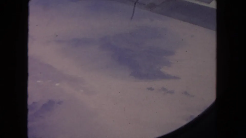Aged 8mm frame from a 1962 airplane-window view over pale desert terrain, with clouds and dark frame edges visible.