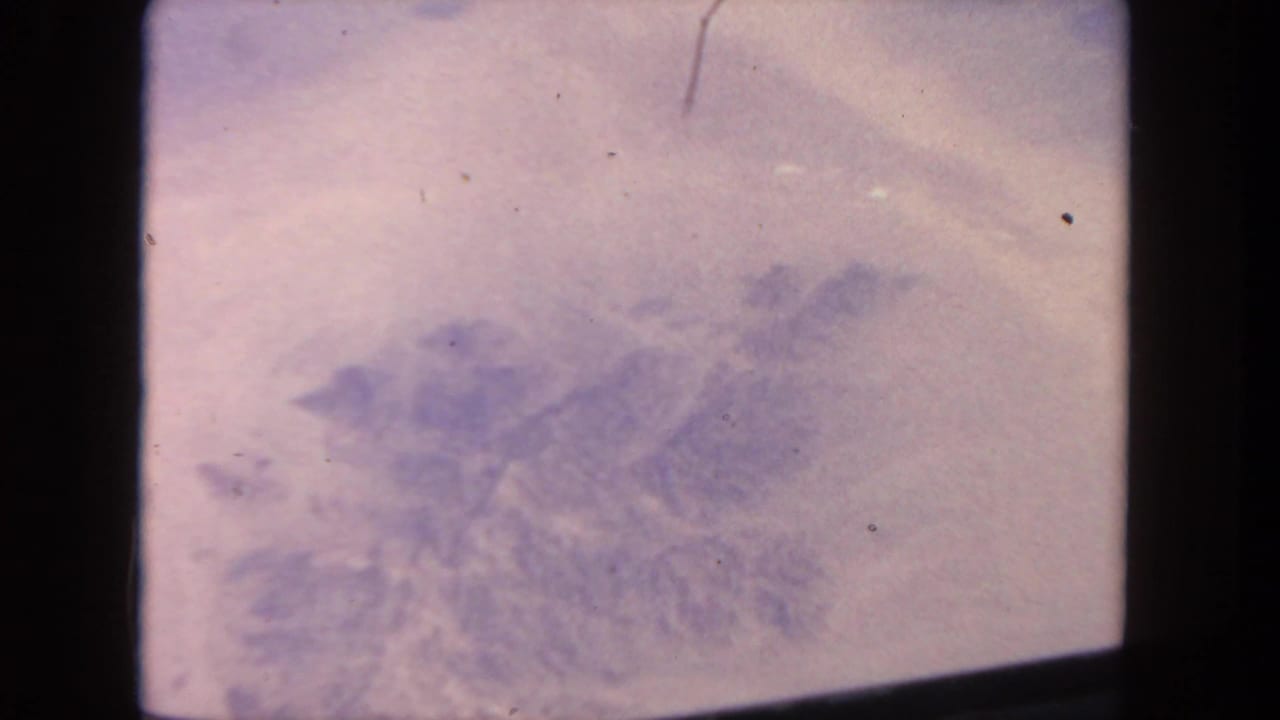 Pale purple aerial view through a dark aircraft-window edge, with indistinct terrain or cloud texture below.