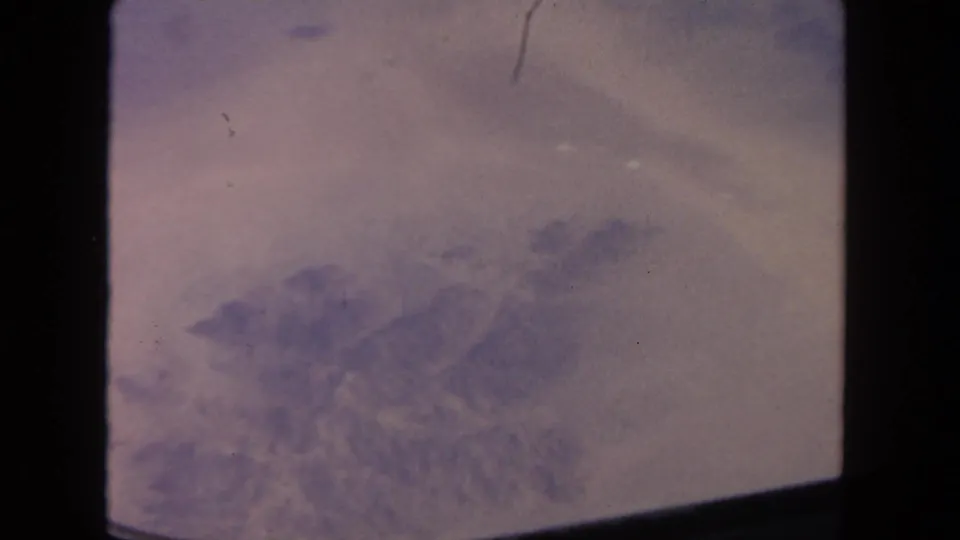 A 1962 home-movie aerial view shows a muted purple landscape or cloud layer beneath the aircraft, bordered by dark film or window edges.