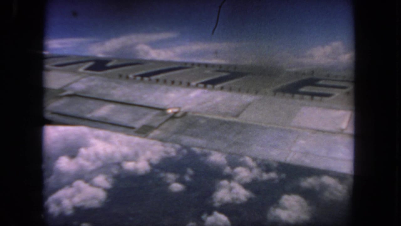 Aircraft wing seen through a passenger window above clouds and blue sky.