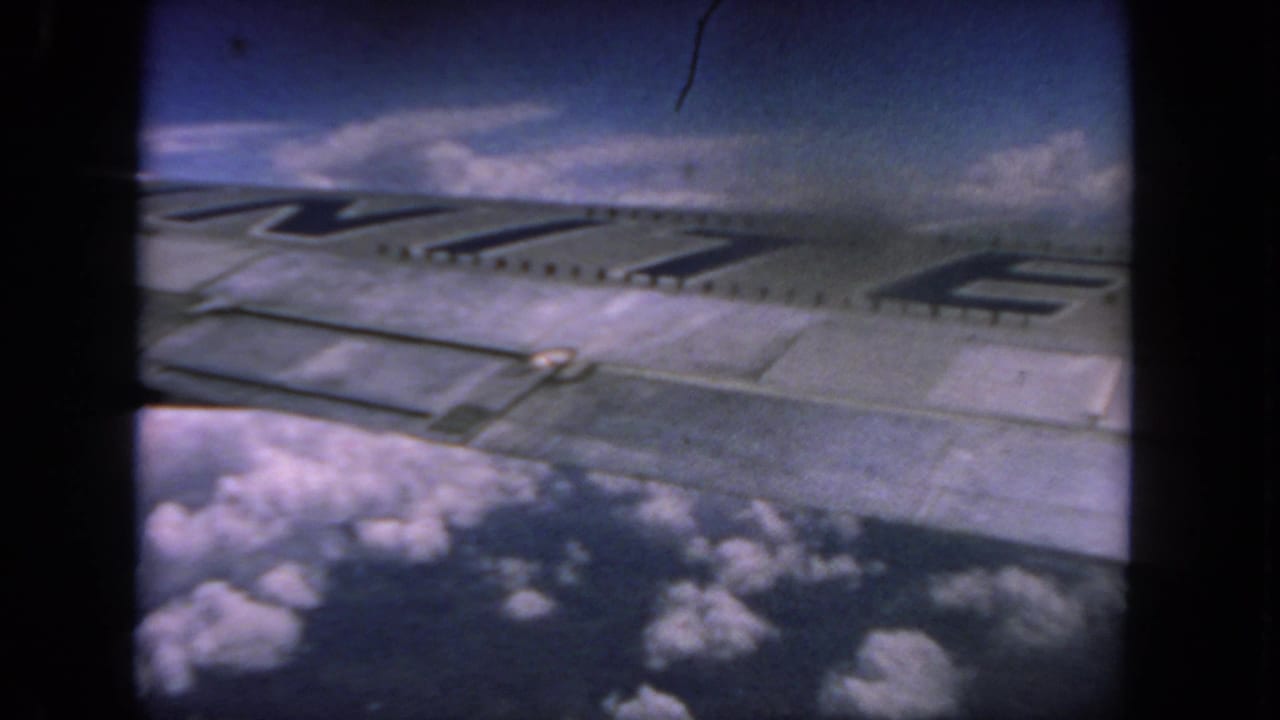 A 1962 airplane-window view shows the wing and partial dark lettering against clouds and a deep blue sky.